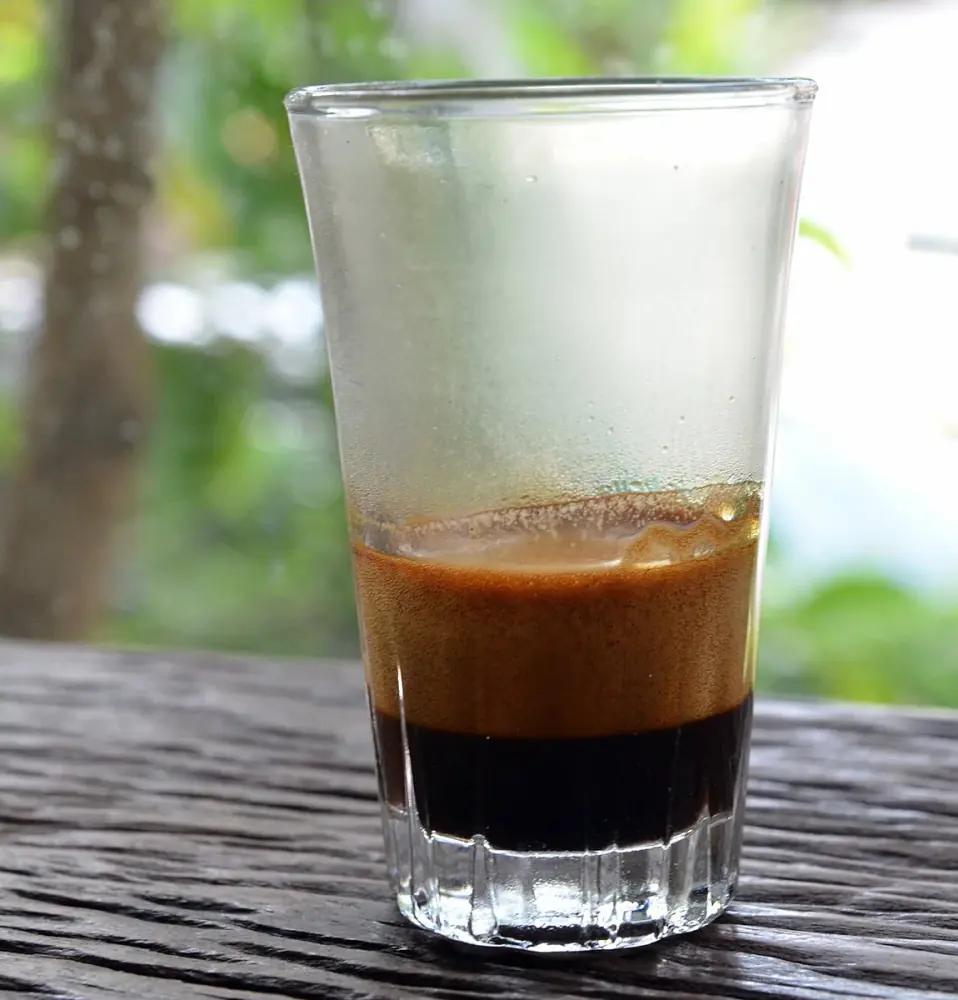 Ly thuỷ tinh đựng espresso, lớp crema nâu vàng nổi trên phần cà phê đen, nền cây xanh mờ