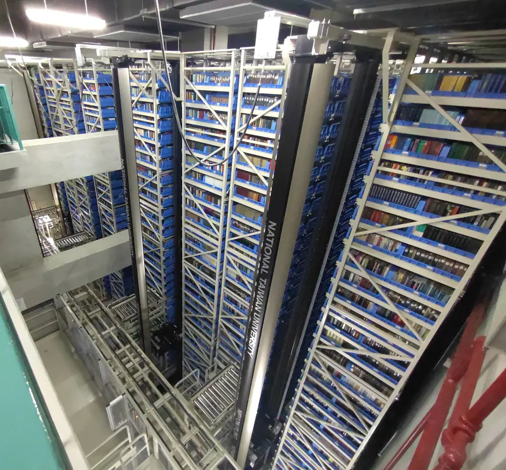 Nhìn từ trên xuống kho tự động: các giàn kệ thép cao nhiều tầng chứa khay xanh, hai cột máy xếp dỡ đen chạy giữa lối đi hẹp