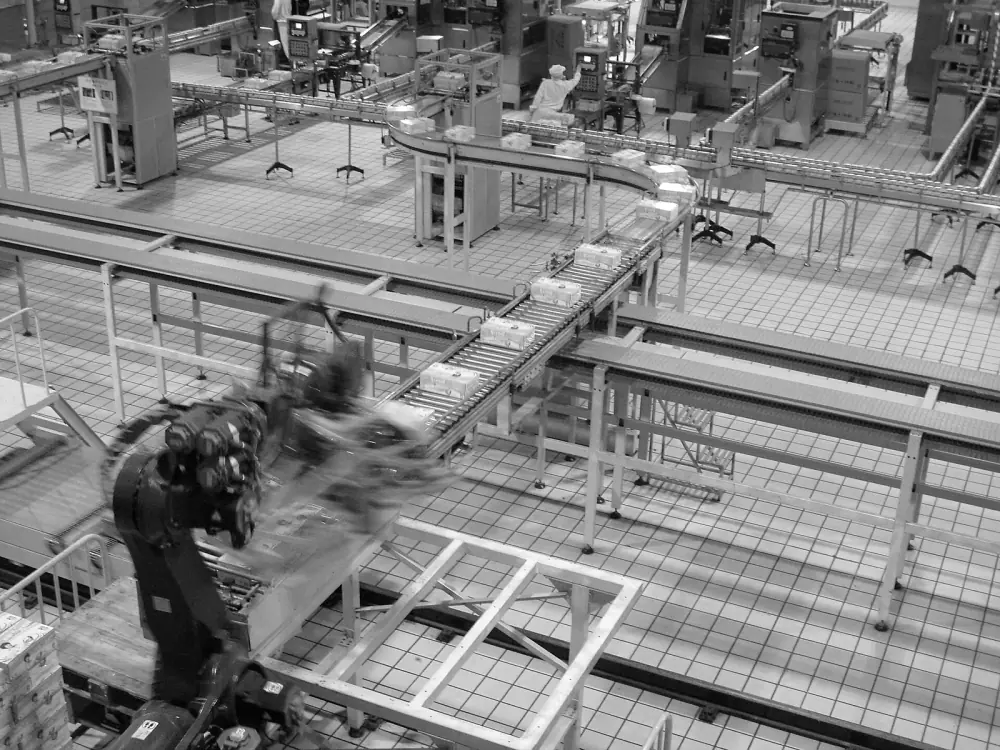Nhìn từ trên cao: robot xếp pallet đang chuyển động mờ ở góc trái, các băng tải con lăn chở hộp hàng chạy ngang nhà máy lát gạch sáng