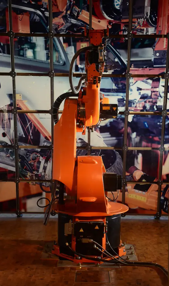 Cánh tay robot công nghiệp màu cam đứng trên bệ đen, các khớp và dây dẫn lộ rõ, phía sau là ảnh lớn dây chuyền lắp ráp ô tô